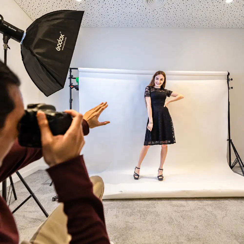 Model in a black dress being photographed on a white backdrop with studio equipment.